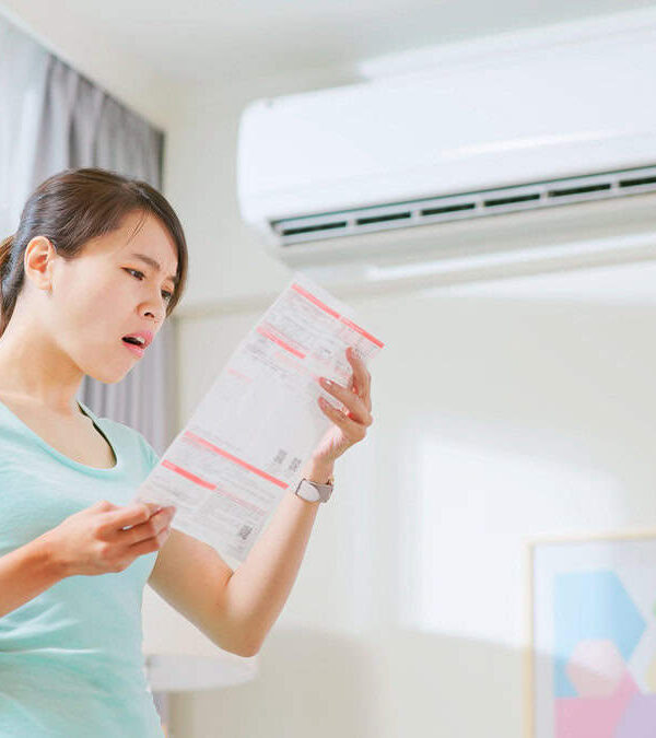 Asian woman stand by air conditioner shocked and upset about electricity bill in hand at home.