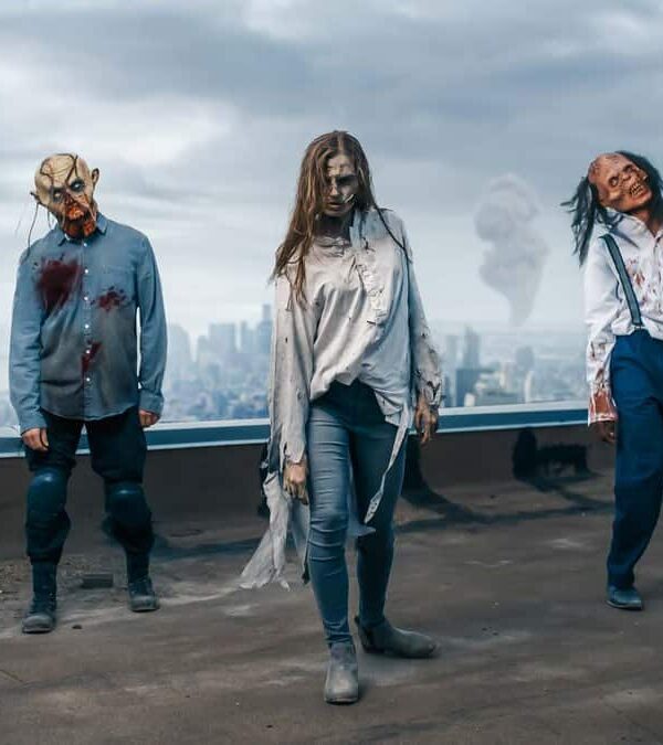 Three people in tattered clothing with zombie makeup stand on a rooftop in a post-apocalyptic cityscape, with smoke rising in the background.