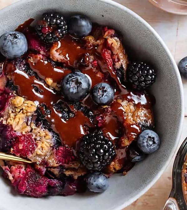 A bowl of baked berry oatmeal topped with fresh blueberries, blackberries, and a drizzle of chocolate sauce, with a gold spoon. Additional blueberries and a baking dish with more oatmeal are nearby.