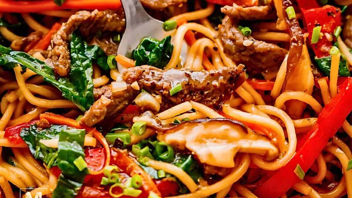 A close-up of a colorful stir-fried noodle dish with beef, red bell peppers, mushrooms, green onions, and leafy greens. A fork is seen in the dish.