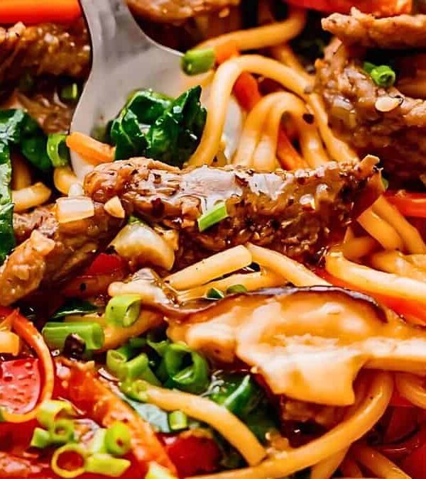 A close-up of a colorful stir-fried noodle dish with beef, red bell peppers, mushrooms, green onions, and leafy greens. A fork is seen in the dish.