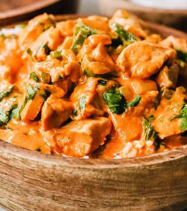 A wooden bowl filled with creamy orange curry containing chicken pieces and garnished with chopped cilantro. Another wooden bowl is partially visible in the background.
