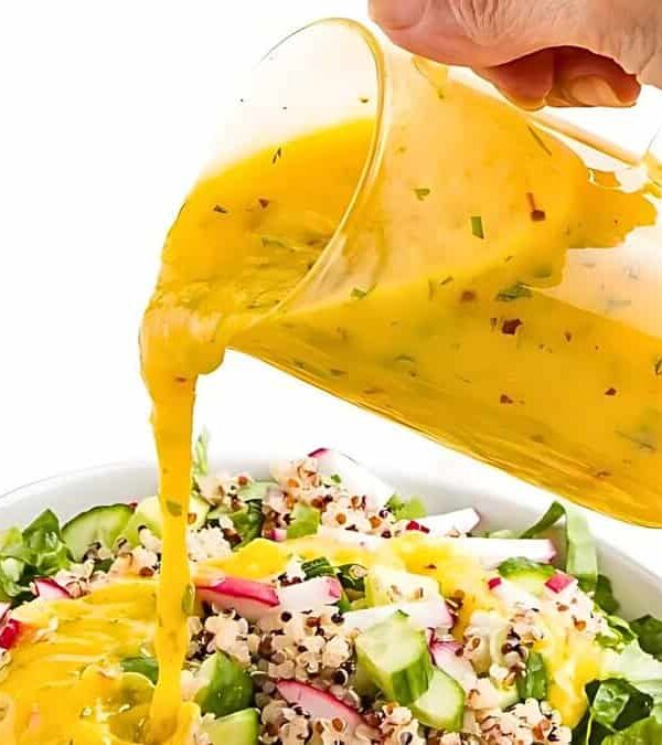 A hand pours orange salad dressing from a glass pitcher onto a bowl of fresh salad containing greens, radishes, quinoa, and other vegetables.