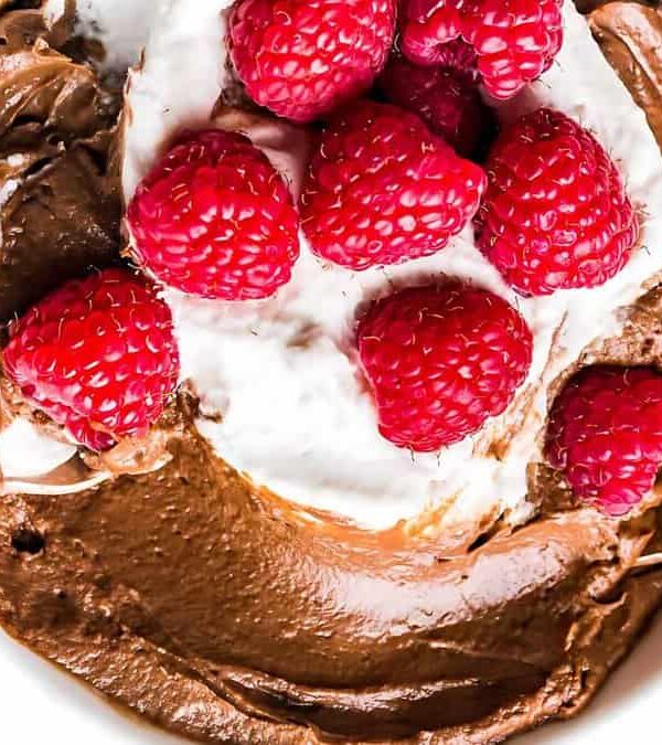 A bowl of chocolate mousse topped with a dollop of whipped cream and fresh raspberries, with two spoons placed inside.