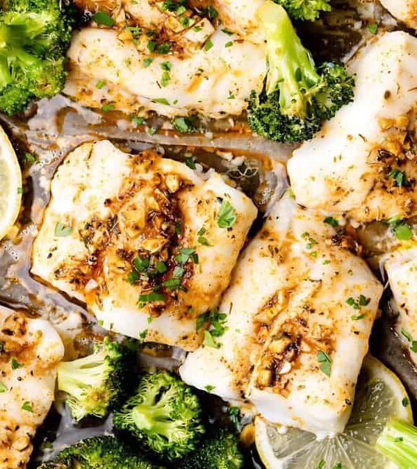 Plated dish featuring baked fish fillets garnished with herbs and spices, surrounded by broccoli florets and lemon slices.