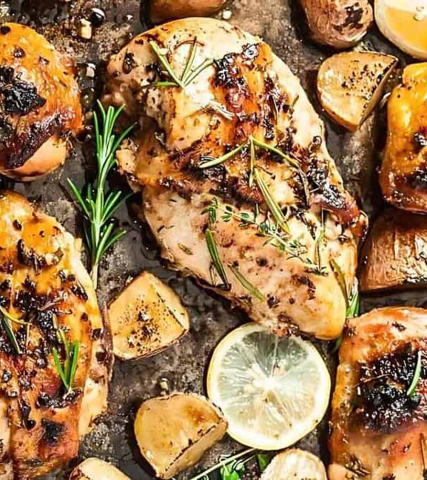 An oven-baked sheet with roasted chicken pieces garnished with lemon slices, rosemary sprigs, and seasoned potatoes.