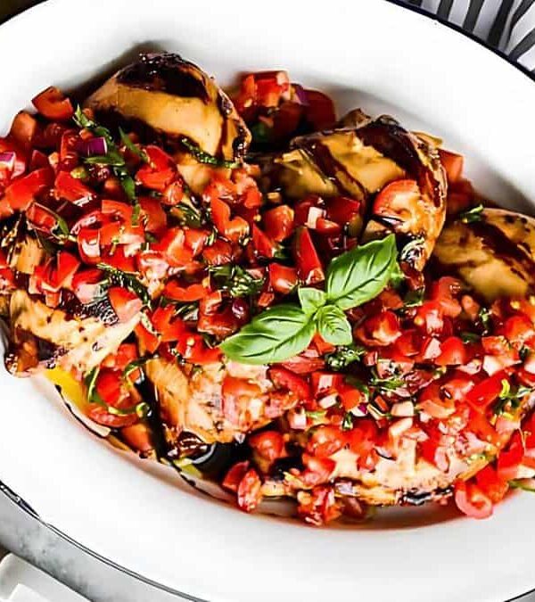 Grilled chicken breasts topped with a tomato, basil, and balsamic vinegar mix, garnished with fresh basil leaves, served on a white oval dish next to whole tomatoes and a striped napkin.