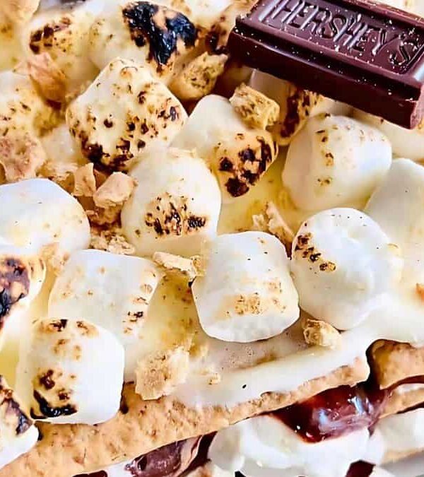 Close-up of a s'mores dessert featuring toasted marshmallows, melted chocolate bars, and graham crackers. Some marshmallows are golden brown, while others are charred.