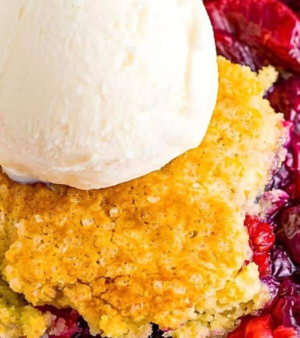 A close-up of a mixed berry cobbler topped with a scoop of vanilla ice cream, featuring a golden brown crust and various berries underneath.