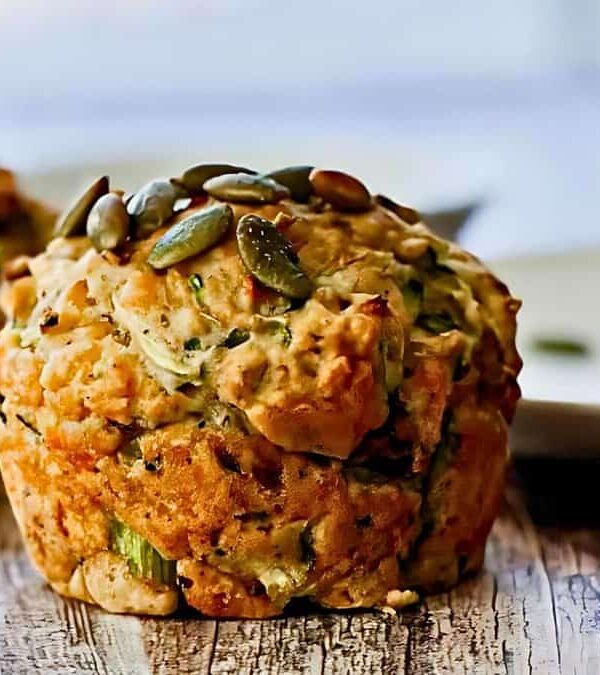 Two zucchini muffins topped with seeds are placed on a wooden surface. One is in focus, while the other is slightly blurred in the background.