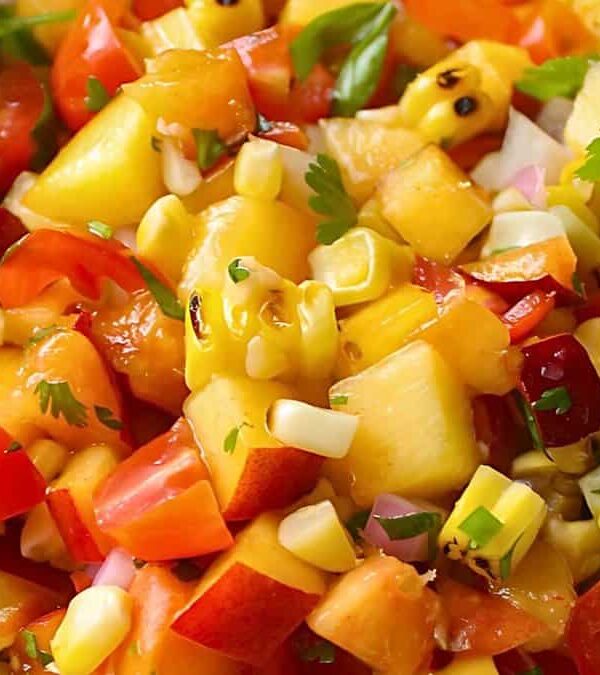 Close-up of a fresh fruit salsa in a bowl, featuring diced peaches, tomatoes, corn, red onions, and cilantro with tortilla chips on the side.