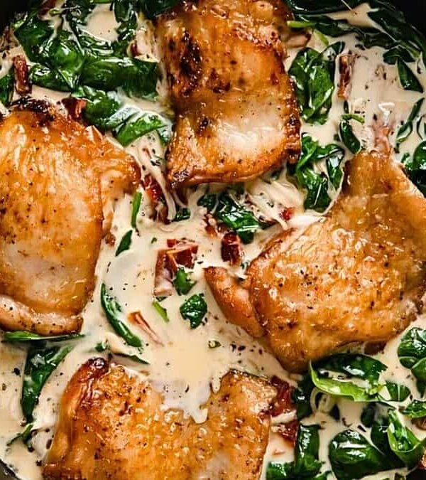 A skillet filled with cooked chicken thighs in a creamy sauce with spinach and sundried tomatoes.