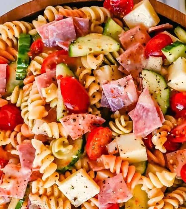 A wooden bowl filled with pasta salad containing rotini, cherry tomatoes, cubed cheese, sliced cucumber, and pieces of salami, seasoned with herbs.