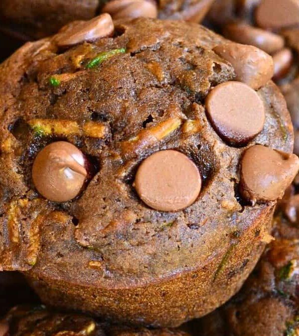 Close-up of several chocolate chip zucchini muffins stacked together. The muffins are topped with chocolate chips and have a moist texture with visible bits of zucchini.