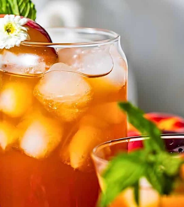 A glass of iced tea garnished with a peach slice, mint leaves, and a small white flower. Another partially visible drink and peaches are in the background.
