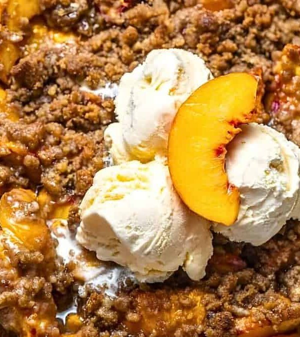 A peach crumble dessert topped with two scoops of vanilla ice cream and a peach slice in a round white dish.