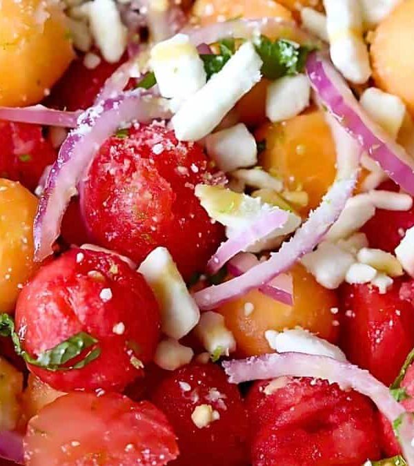 Close-up of a colorful salad featuring balls of watermelon and cantaloupe, sliced red onions, crumbled feta cheese, and chopped mint leaves.