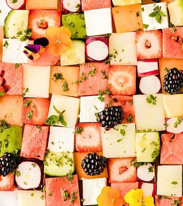 A colorful fruit and vegetable platter arranged in a checkerboard pattern, featuring cubes of watermelon, cucumber, and other produce, garnished with blackberries and edible flowers.