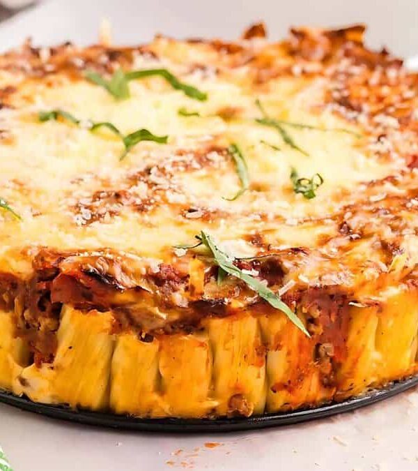 A baked pasta pie with cheese and tomato sauce, garnished with basil, on a white plate. A serving utensil and a small bowl of greens are nearby.