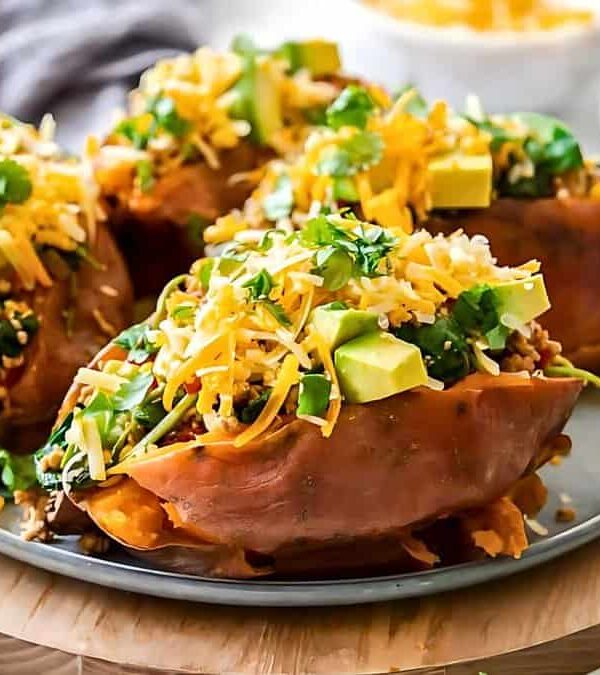 Three baked sweet potatoes topped with avocado, shredded cheese, cilantro, and other garnishes, served on a round plate with a wooden base. Additional toppings in bowls are in the background.