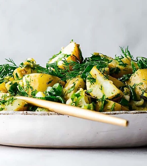 A white plate filled with a potato salad, garnished with fresh herbs such as dill and mixed greens, with a gold spoon resting on the side.