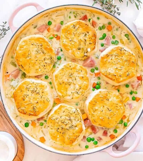 A round dish filled with creamy chicken pot pie, topped with seven golden biscuits, and garnished with herbs. Peas and carrots are visible. A bowl of mixed peas and carrots, garlic, and spices are nearby.