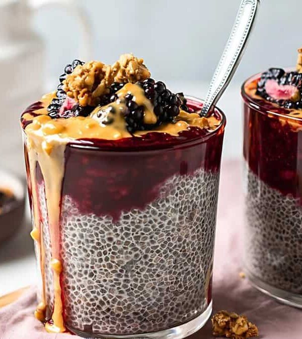 Two glasses filled with chia pudding topped with blackberries, granola, and a drizzle of nut butter, with spoons inside each glass. A plate with granola is in the background.