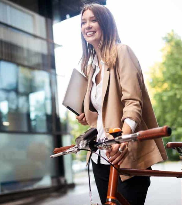 A person in business attire smiles while walking their bicycle outside a glass building, holding a laptop.