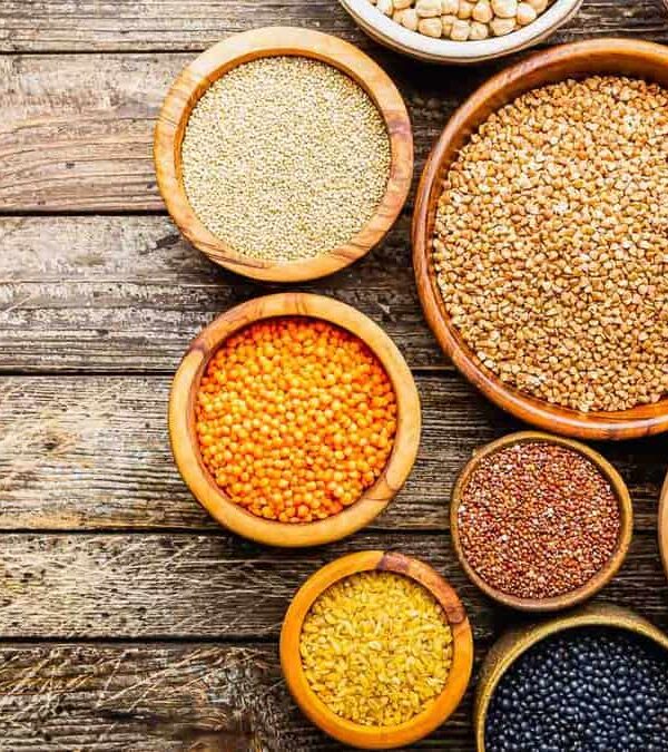 Various grains and legumes displayed in wooden bowls on a rustic wooden surface.