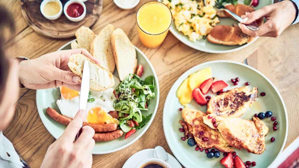 Two people eating a breakfast spread that includes toast, eggs, sausages, a salad, fruit-topped French toast, a glass of orange juice, coffee, and assorted sauces.