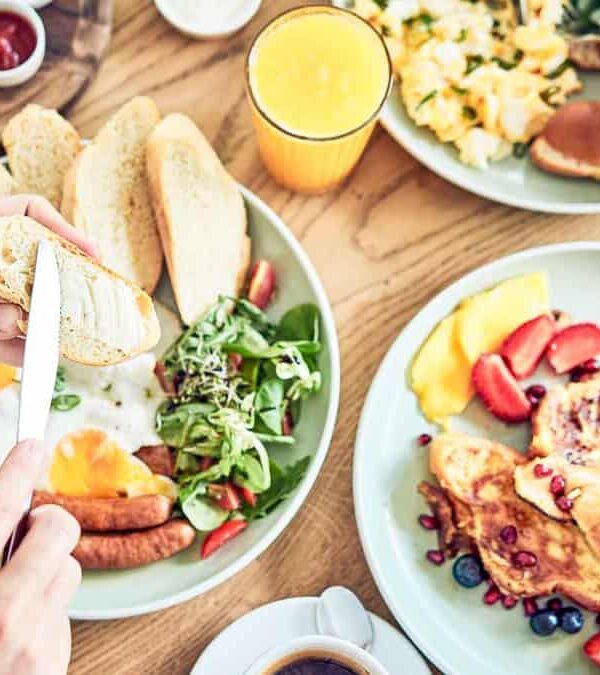 Two people eating a breakfast spread that includes toast, eggs, sausages, a salad, fruit-topped French toast, a glass of orange juice, coffee, and assorted sauces.