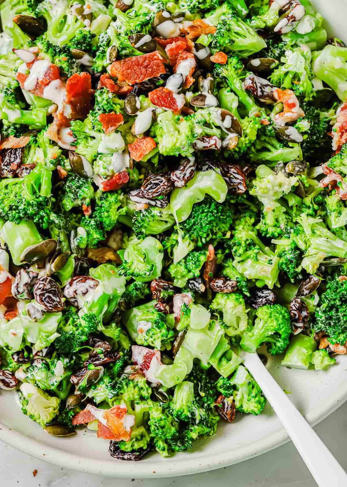 A close-up of a broccoli raisin salad mixed with bacon bits and seeds served on a white plate with a white utensil visible to the side.