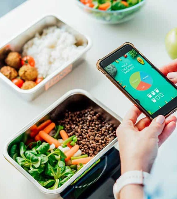 Close-up of a person holding a smartphone showing a diet tracking app, with a table in the background featuring meal prep containers with rice, vegetables, meatballs, and seasoning items.