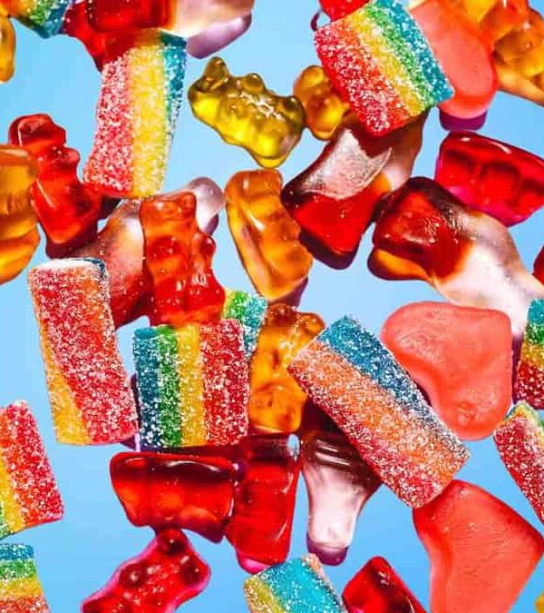 A variety of colorful gummy candies, including gummy bears, rainbow strips, and heart shapes, are scattered against a blue background.