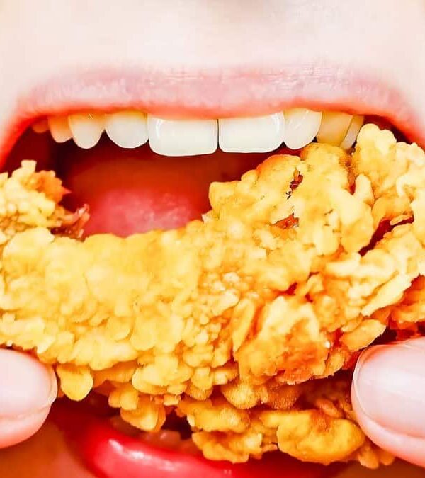 Close-up of a person eating a piece of crispy fried chicken, showing their teeth and fingers gripping the chicken.
.