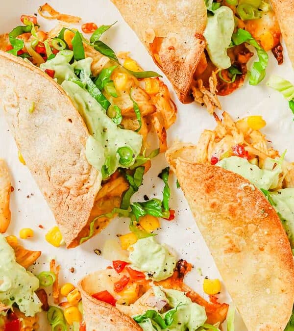 A tray of fried chicken tacos filled with shredded chicken, diced vegetables, and topped with a creamy green sauce and chopped green onions.