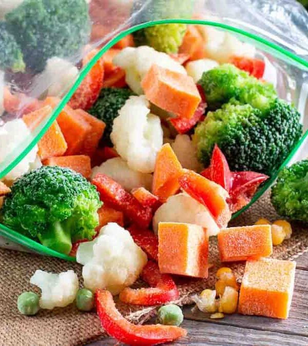A plastic bag lies open, spilling out a mix of frozen vegetables including broccoli, cauliflower, carrots, red peppers, and peas, onto a burlap mat on a wooden surface.