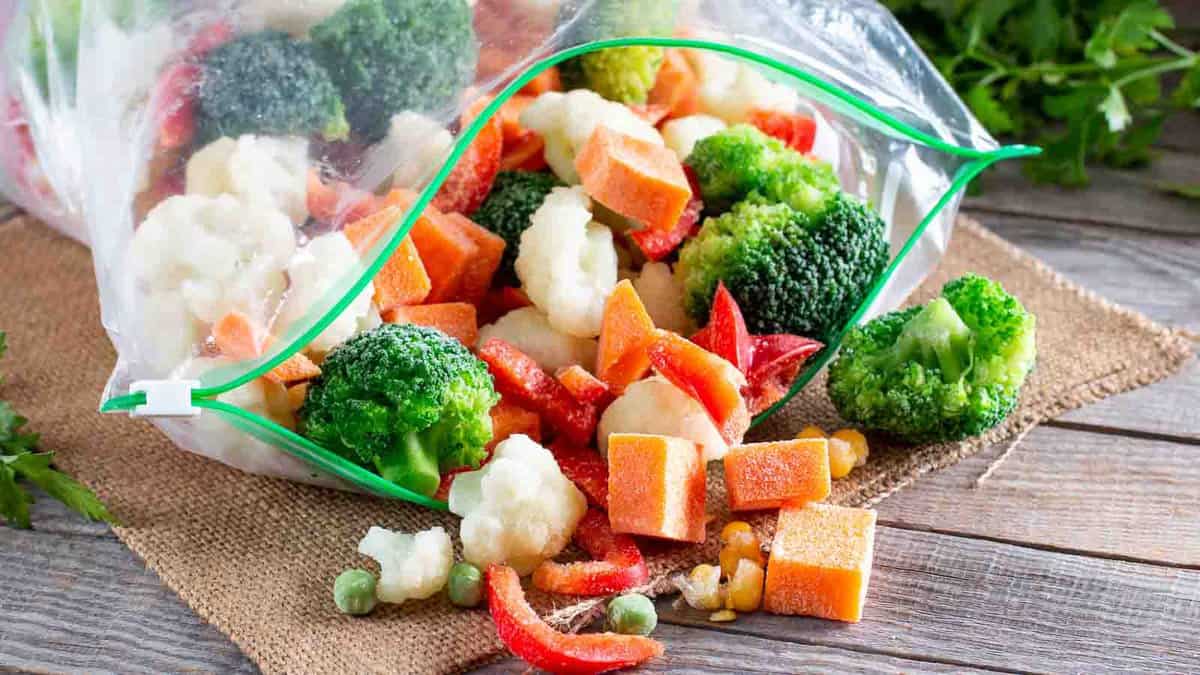 A plastic bag lies open, spilling out a mix of frozen vegetables including broccoli, cauliflower, carrots, red peppers, and peas, onto a burlap mat on a wooden surface.