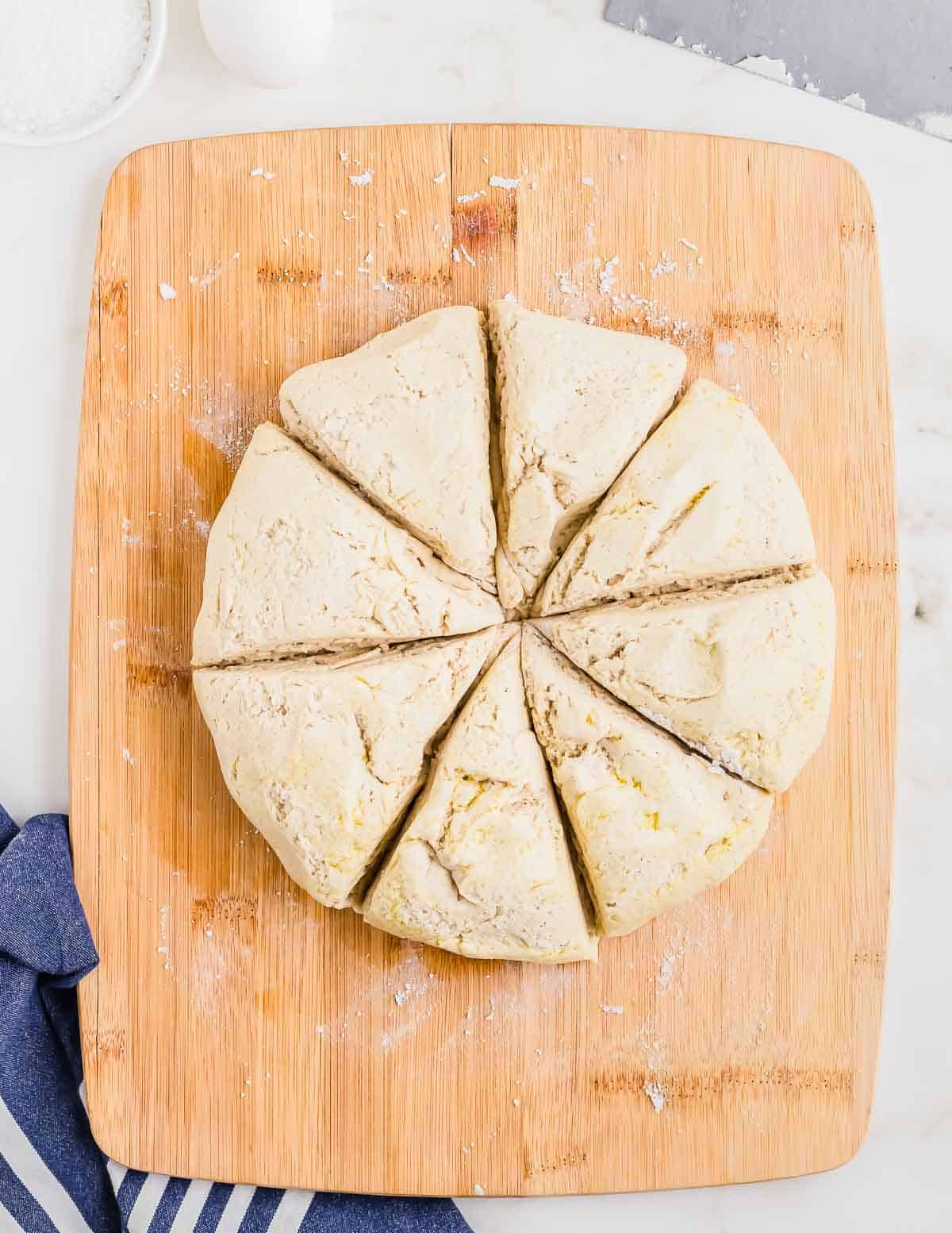 A round piece of dough cut into eight triangular sections on a wooden cutting board, with a blue and white striped cloth partially visible in the lower left corner.