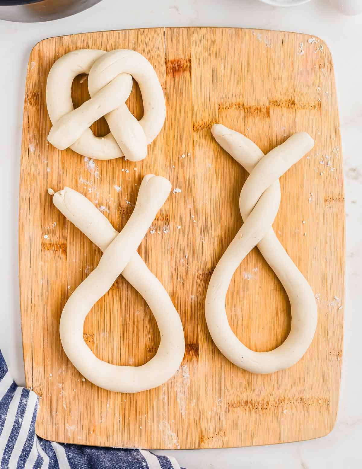 Three pretzel dough shapes placed on a wooden cutting board. The top left one resembles a traditional pretzel shape, while the other two are partially twisted into an elongated form.