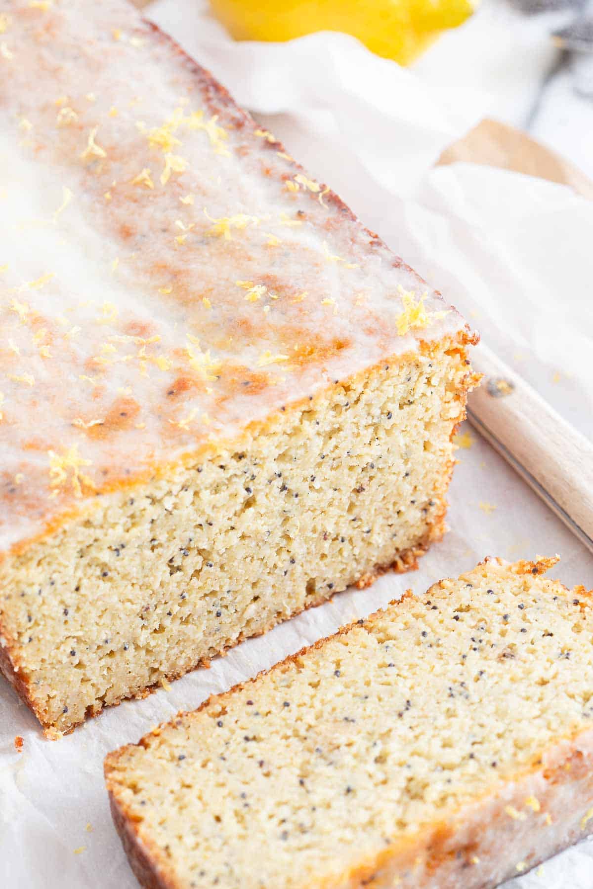 A freshly sliced loaf of lemon poppy seed bread rests on parchment paper. The bread has a light glaze on top and visible poppy seeds throughout.