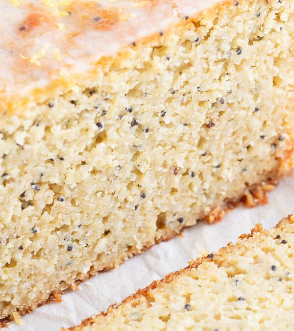 A close-up of a loaf of lemon poppy seed bread, with a few slices cut from it. The bread has a golden-brown crust and visible poppy seeds scattered throughout.