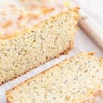 A loaf of lemon poppy seed cake with one slice cut and placed on a white parchment paper surface. The cake has a visible glaze on top and is dotted with poppy seeds.
