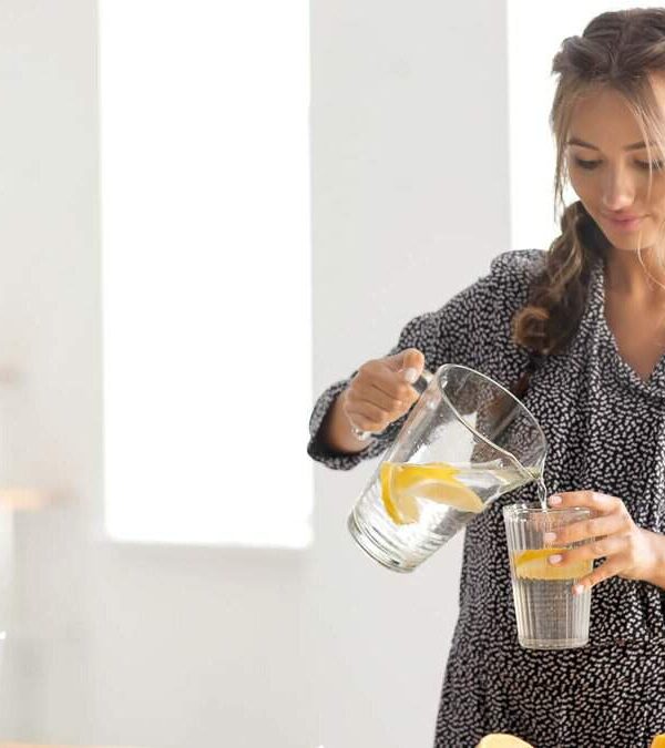 Woman in a patterned dress pouring lemon water from a glass pitcher into a glass in a bright indoor setting.