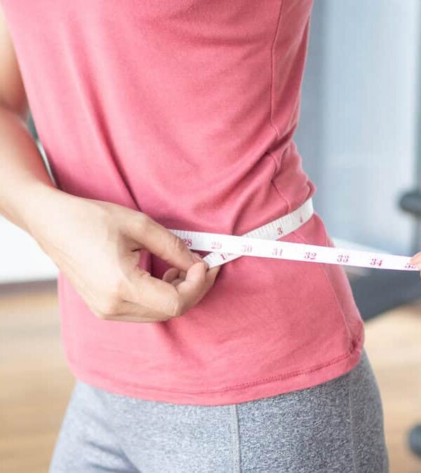 Person in a pink shirt and gray pants measuring their waist with a tape measure in a gym setting.