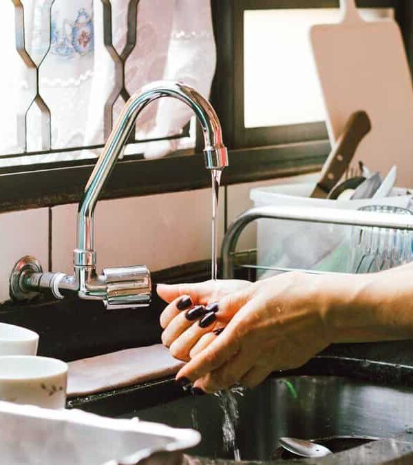 A person is washing their hands under a kitchen sink faucet. Various kitchen items, including cups and a pitcher, are visible on the counter nearby.