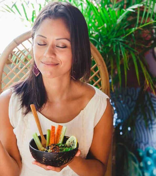 A woman in a white top sits in a wicker chair, holding a bowl of vegetables and a dip. She has her eyes closed and smiles slightly, with her head tilted slightly to one side. Plants are in the background.