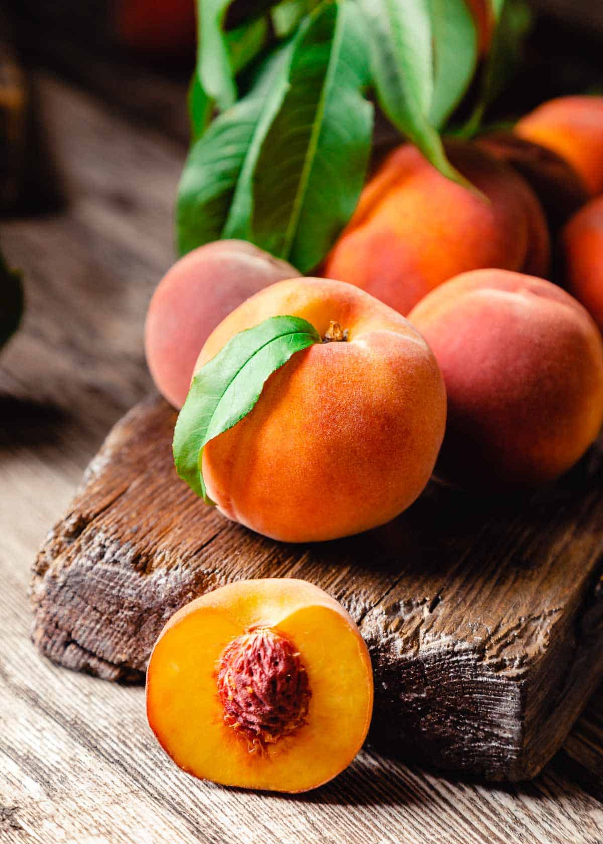Cluster of fresh peaches with green leaves on a wooden surface, one peach cut open revealing the pit.