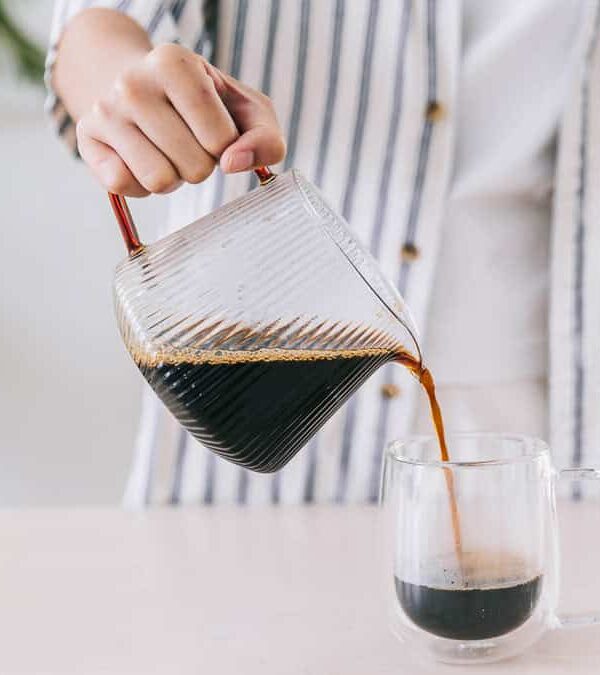 A person pours coffee from a glass carafe into a clear glass mug on a cream-colored countertop. They wear a striped shirt, and a leafy plant is in the background.