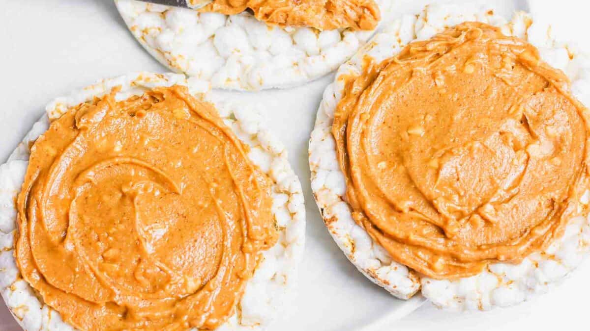 Three rice cakes spread with peanut butter are arranged on a white surface. One additional rice cake is in the process of being spread with peanut butter.